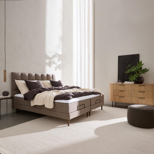 Lyst, minimalistisk soveværelse med polstret seng, beige tekstiler, trækommode med grøn plante og højt vindueslys.