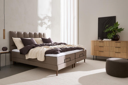 Lyst, minimalistisk soveværelse med polstret seng, beige tekstiler, trækommode med grøn plante og højt vindueslys.