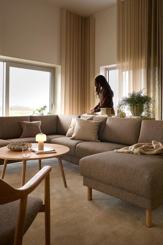 Lyst stue med beige gardiner, brun hjørnesofa i stof, rundt træbord med lille lampe og plaid på chaiselongen; en person står ved vinduet.