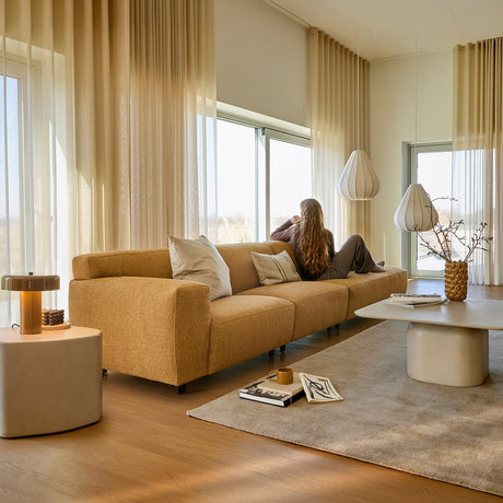 Lyst stue med beige, lette gardiner, sennepsgul sofa med puder, to hvide pendellamper og lavt bord med vase og grene på et stort tæppe; en person sidder ved vinduet og kigger ud.