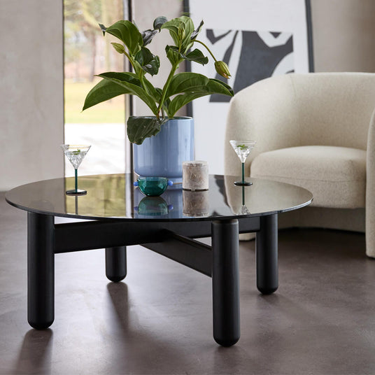 Sort rundt sofabord med glasplade, beige lænestol, blå vase med grøn plante og to martiniglas i en lys moderne stue.