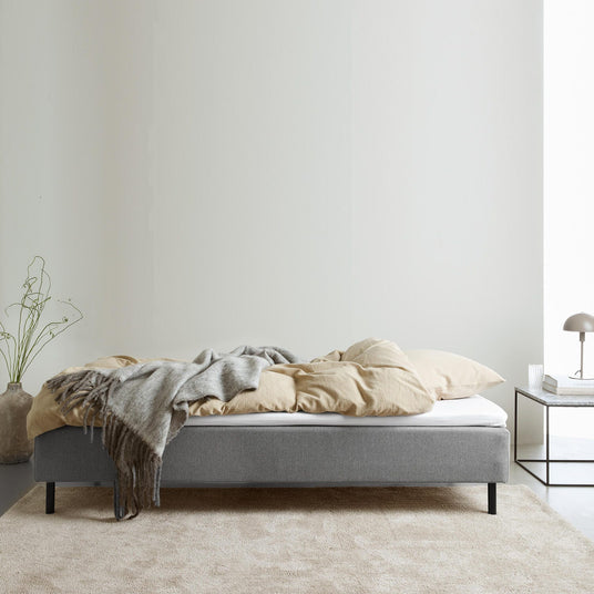Grå polstret seng med beige sengetøj og grå plaid; natbord med lampe og bøger i et lyst, minimalistisk soveværelse.