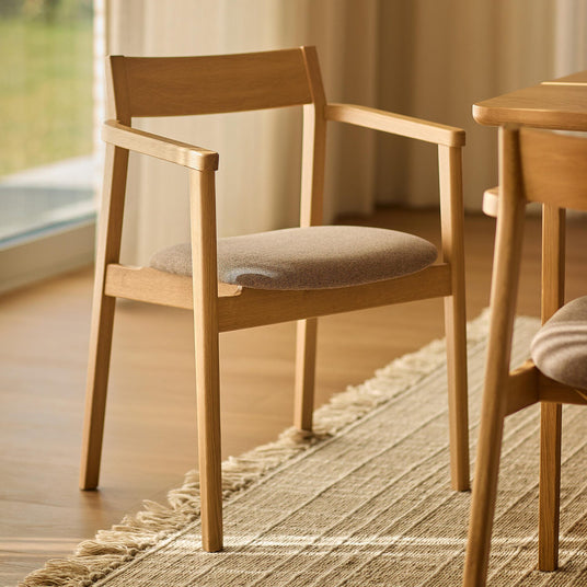 Lys egetræs spisebordsstol med armlæn og beige stofsæde på en jutelignende måtte i et lyst spiserum.