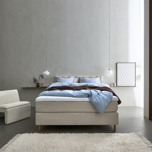 Minimalistisk soveværelse med beige kontinentalseng, lyseblåt sengetøj, to hvide pendellamper og et blødt lyst tæppe i roligt dagslys.