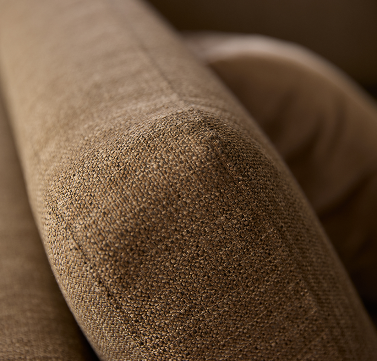 Nærbillede af sofaens armlæn i brun vævet tekstil med blødt afrundet form og en pude i baggrunden.