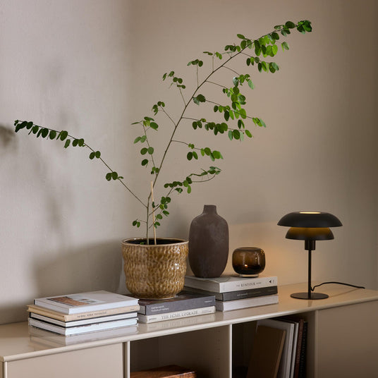 Beigefarvet skænk med grøn potteplante, keramikvase, tændt lysglas og sort bordlampe oven på stakke af bøger som The Kinfolk Home og The Home Upgrade mod en neutral væg.