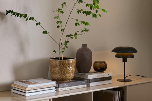 Beigefarvet skænk med grøn potteplante, keramikvase, tændt lysglas og sort bordlampe oven på stakke af bøger som The Kinfolk Home og The Home Upgrade mod en neutral væg.