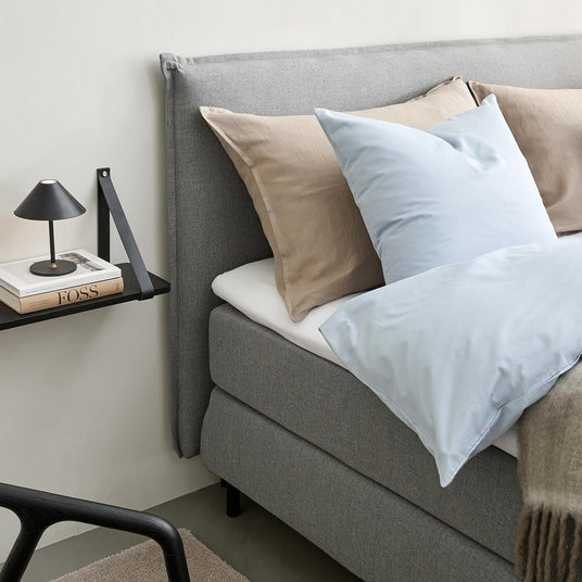 Grå polstret seng med beige og lyseblå puder, plaid og væghylde med sort lampe og bøger inklusiv FOSS.