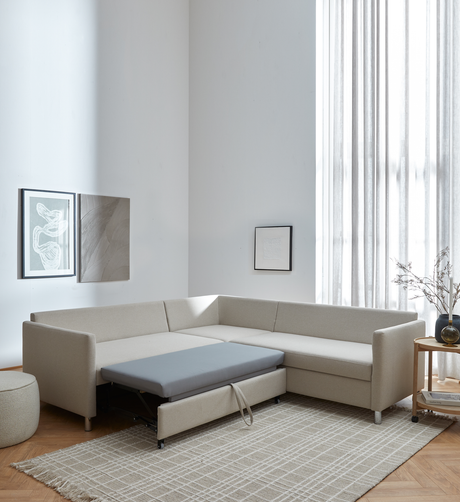 Beige L-formet hjørnesofa med udtræksseng trukket frem i en lys, minimalistisk stue med lette gardiner, neutral ternet tæppe, vægkunst og rundt sidebord med vase.