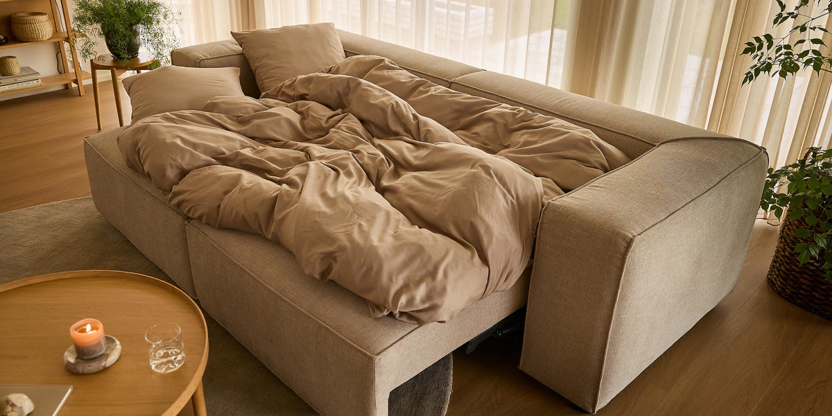 Beige sovesofa slået ud med sandfarvet sengetøj i en lys stue med lette gardiner, rundt træbord med tændt lys og grønne planter.