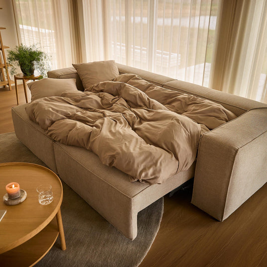 Beige sovesofa slået ud med sandfarvet sengetøj i en lys stue med lette gardiner, rundt træbord med tændt lys og grønne planter.