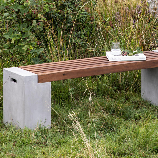 Udendørs bænk med betonben og trælameller, med et glas vand og bogen ”barefoot living” ovenpå i græsset.