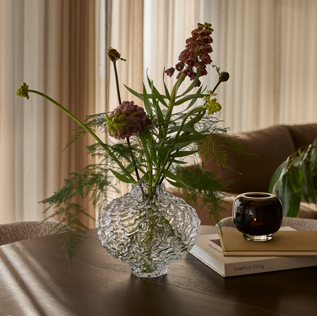 Struktureret glasvase med blomster på et træbord ved siden af en brun lyslygte på to bøger, hvor den nederste har titlen ”barefoot living”, i blødt dagslys med gardiner i baggrunden.