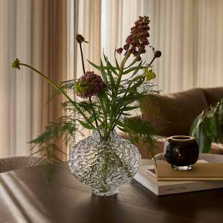 Bølgestruktur glasvase med blomster på træbord ved sofa; rund mørk lyslygte på bøger, herunder ”barefoot living”, bløde gardiner i baggrunden.