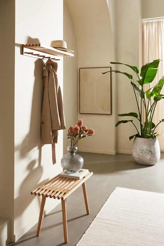 Lys entré med træbænk, vægmonteret hylde med beige frakke og stråhat, stor grøn plante og glasvase med blomster.