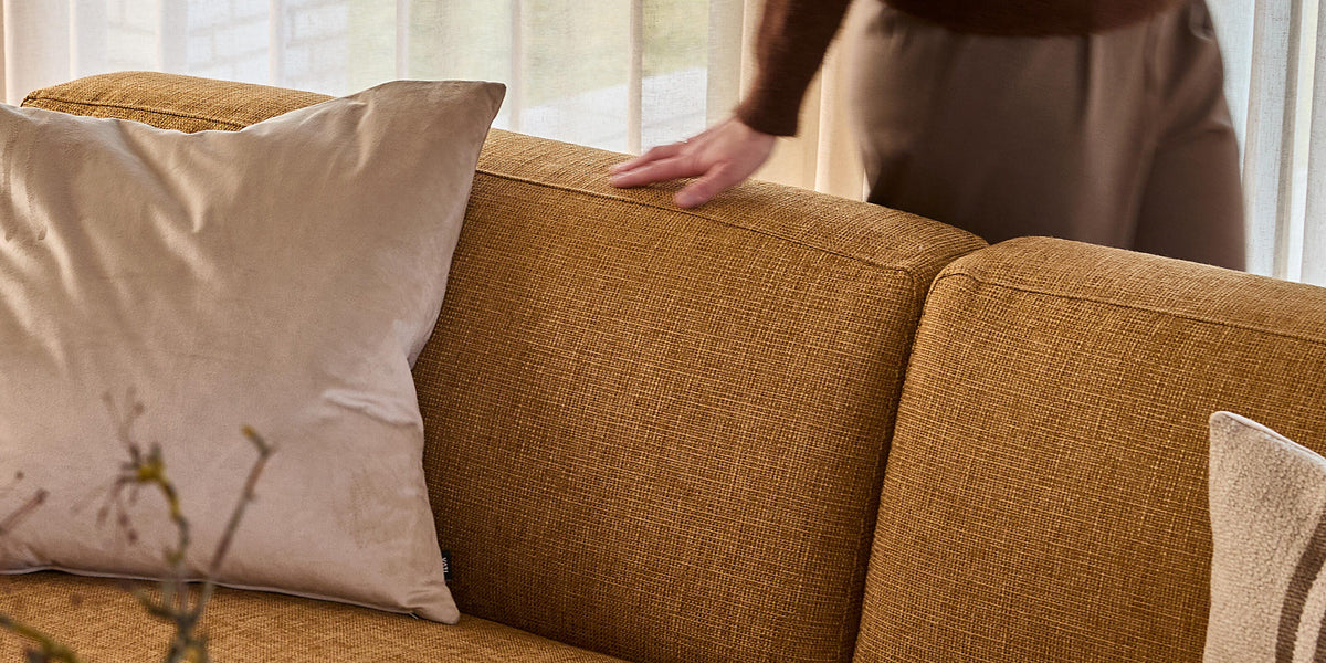 Okker-gul tekstilsofa med beige puder; en hånd hviler på ryglænet foran lette gardiner i dagslys.