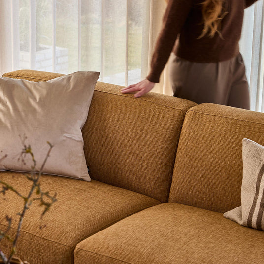 Okker-gul tekstilsofa med beige puder; en hånd hviler på ryglænet foran lette gardiner i dagslys.