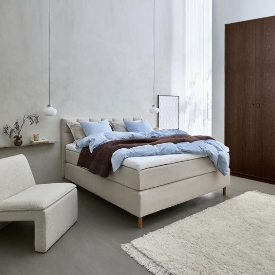 Lyst skandinavisk soveværelse med beige polstret seng, lyseblåt sengetøj og brun plaid, to pendler, lav lounge-stol, vase med grene på hylde, lette gardiner, hvid langhåret tæppe og mørkt klædeskab.