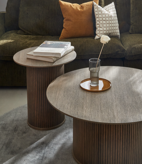 To runde sofaborde i mørk eg med riflede sider; på det ene ligger bøger, herunder ”Nordic Interiors”, på det andet står en glasvase med en hvid blomst på et brunt fad foran en olivengrøn sofa med rustfarvet og hvidternet pude.