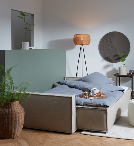 Lyst stue med sovesofa redt med blågråt sengetøj, morgenmadsbakke med tekande, flettet gulvlampe, grønne planter og rund vægdekoration.