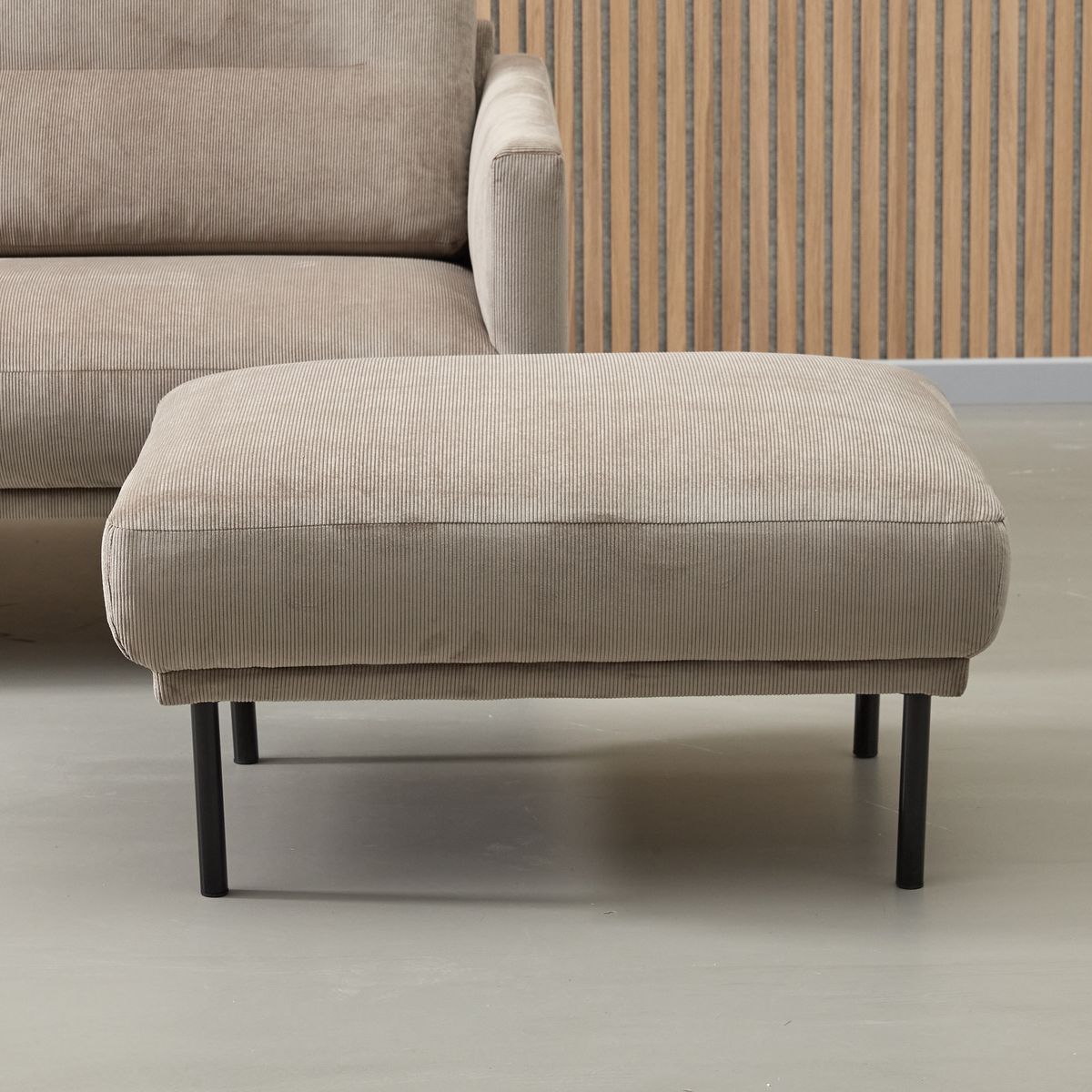 Beige puf i fløjl med sorte metalben i stue, matchende sofa og lameltrævæg.
