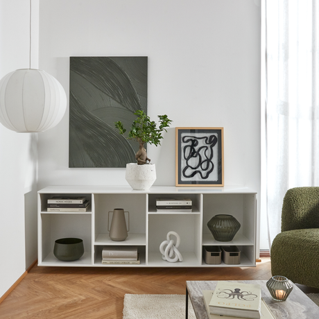 Minimalistisk stue med hvid opbevaringsreol, abstrakt vægkunst, bonsai i krukke og rund pendellampe ved siden af grøn sofa i dagslys.