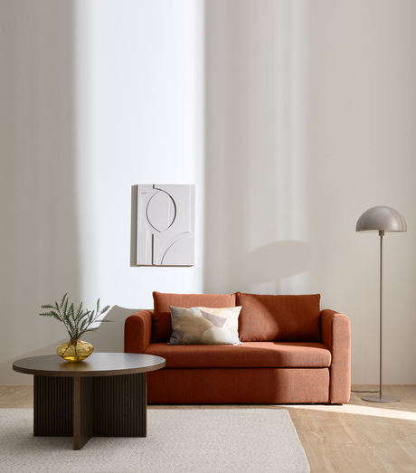 Lyst stue med terrakottafarvet sofa, lys tæppe, mørkt rundt sofabord med gul glasvase og grøn gren, minimalistisk vægkunst og en beige gulvlampe i blødt dagslys.