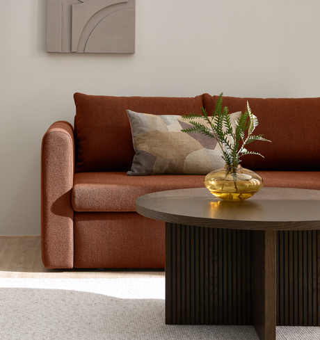 Rustorange sofa med mønstret pude, mørkt rundt sofabord og gul glasvase med grønne kviste i en lys stue.