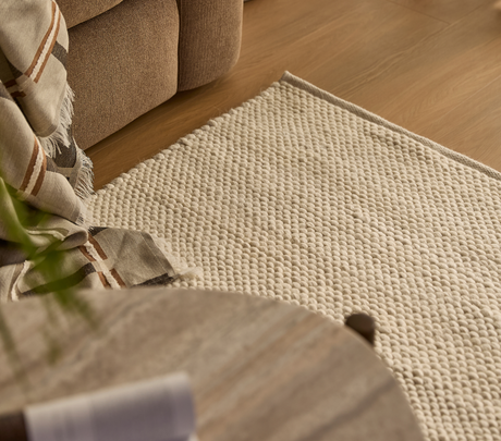 Nærbillede af beige, tekstureret tæppe ved siden af sofa med ternet plaid på trægulv i en hyggelig stue.