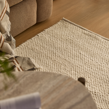 Nærbillede af lys, struktureret tæppe ved siden af beige sofa, med stribet plaid og trægulv i varmt dagslys.