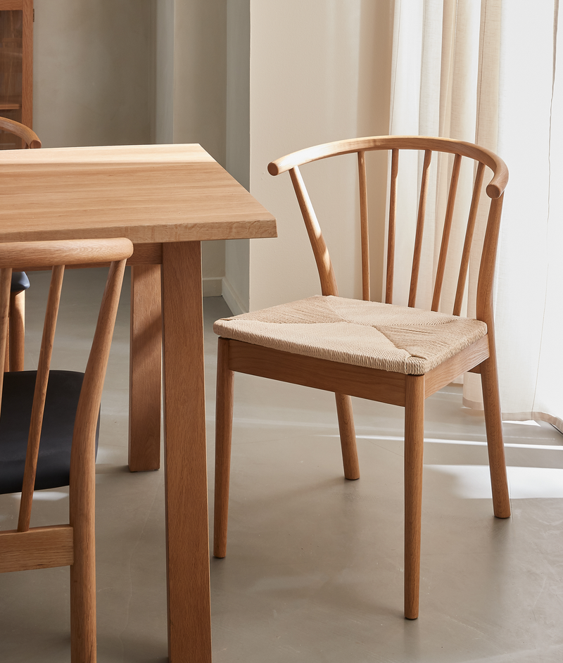 Træstol med buet ryg og fletsæde ved siden af et egetræsbord i et lyst, minimalistisk spisemiljø.