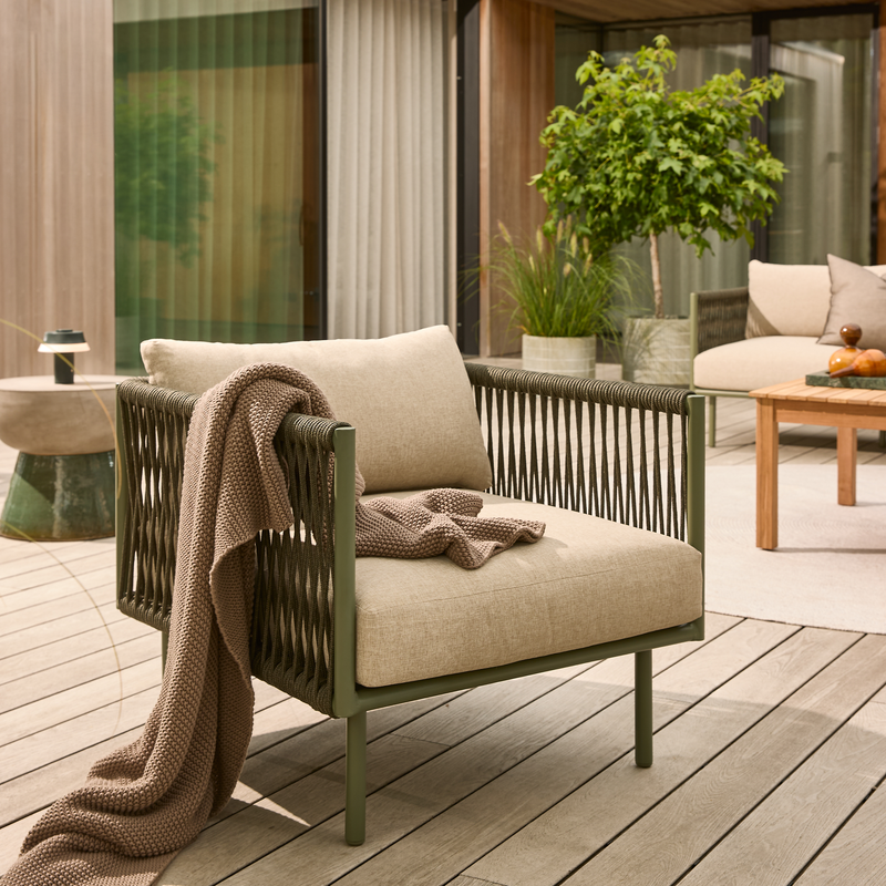 Udendørs lænestol med beige hynder og en strikket plaid på en træterrasse, omgivet af potteplanter og et lavt bord i en moderne patio.
