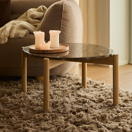 Lavt sofabord i træ med mørk marmorplade på lurvet beige tæppe, bakke med tændte lys ved siden af sofa med plaid.