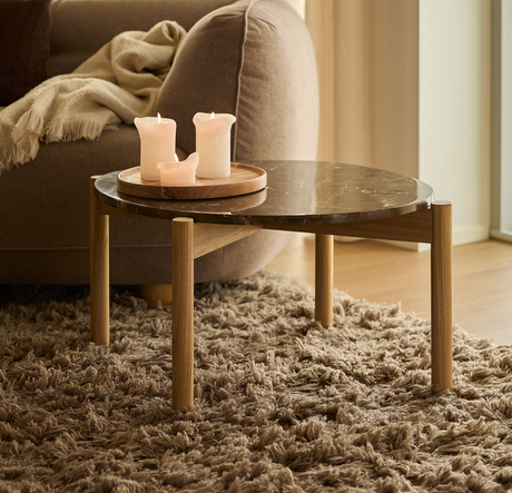 Lavt sofabord med mørk marmorlignende bordplade og træben på brun højluvet tæppe, med tændte lys på en rund træbakke ved en beige sofa i dagslys.