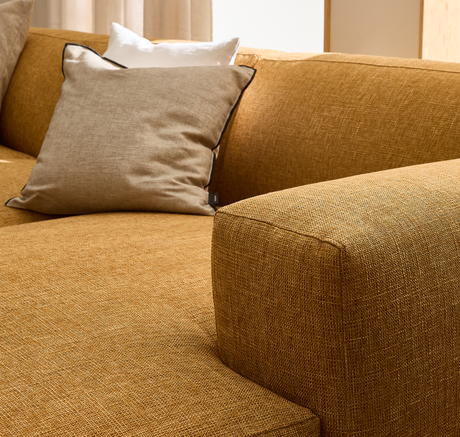 Nærbillede af en sennepsgul stofsofa med en beige pude med mørk kant og en hvid pude bagved i et lyst stue-miljø.