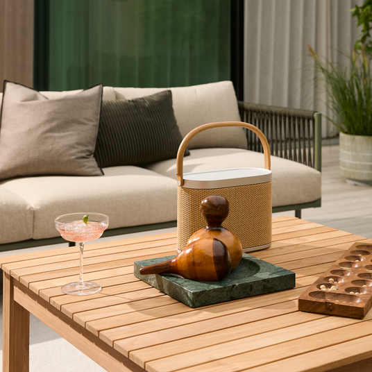 Træbord på terrasse med cocktail i coupeglas, bærbar højttaler med hank, træskulptur på grøn stenplade og mancala-spil; beige udendørs sofa og plante i baggrunden.
