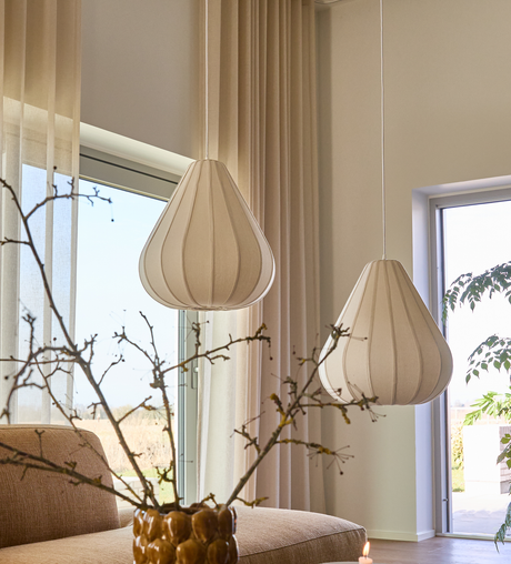 To beige stofpendler over sofa ved store vinduer, lette gardiner, vase med grene og dagslys.