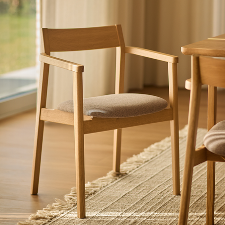 Lys træstol med armlæn og beige sæde ved spisebord på lys vævet tæppe i solfyldt rum.