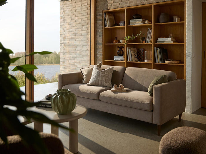 Lys skandinavisk stue med beige sofa, indbygget reol i træ, rundt sofabord med grøn vase og store vinduer med udsigt over en sø.