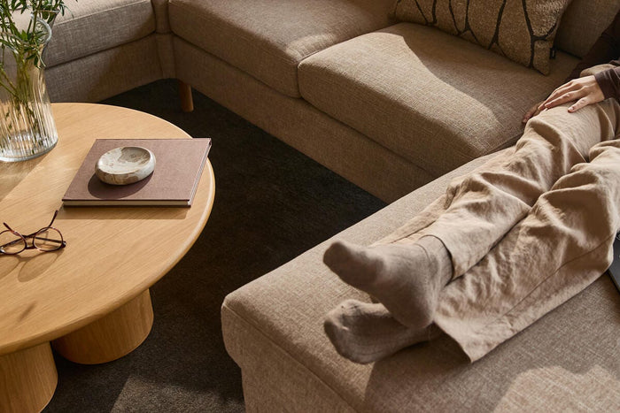 Beige hjørnesofa med puder; person med sokker hviler benene på chaiselongen. Rundt sofabord i træ med vase, briller og bog i en varm, neutral stue.