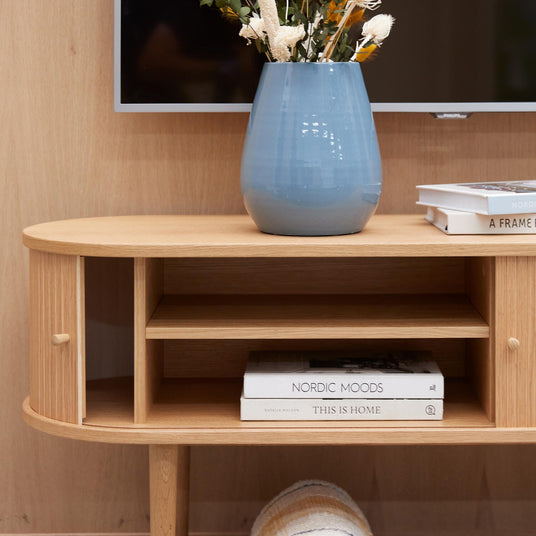 Lyst træmediemøbel med blå vase med tørrede blomster; bøgerne “NORDIC MOODS” og “THIS IS HOME” ligger på hylden og et TV ses ovenover.