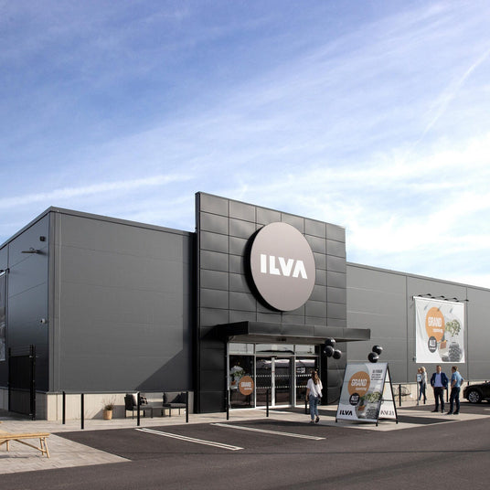 ILVA varehus set udefra med indgang og skilte for Grand Opening, mennesker og biler foran under blå himmel.