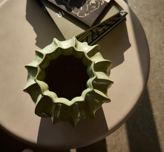 Grøn skulpturel vase set oppefra på et rundt bord med et par bøger i varmt sollys.