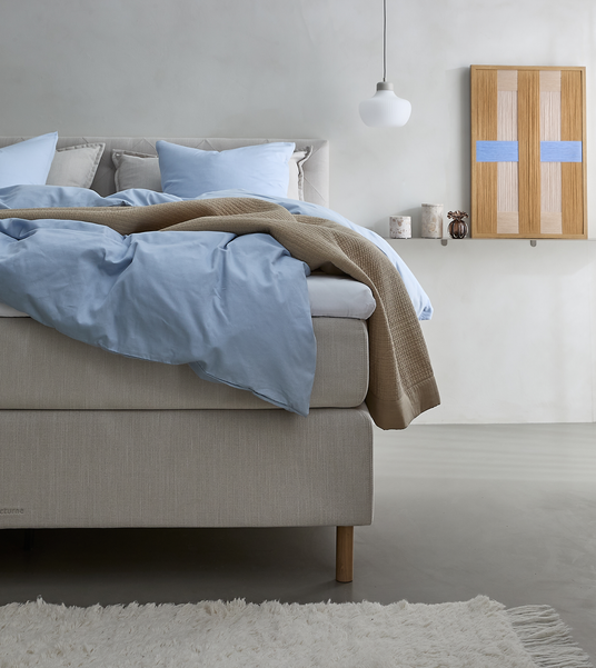 Lyst soveværelse med beige seng, lyseblåt sengetøj, beige plaid, hvide puder, hængende pendellampe, vægkunst og stearinlys på en lille hylde; blød hvid måtte på gulvet.