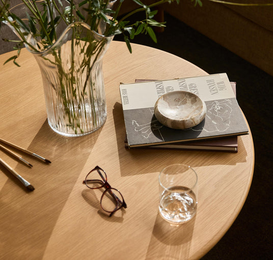 Rundt sofabord i lyst træ med glasvase med grønne grene, en stak bøger med en lille stenskål ovenpå, briller, pensler og et glas vand i blødt dagslys.