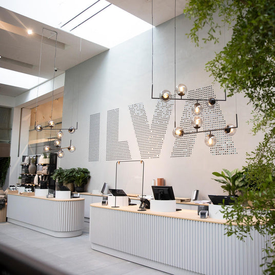 Moderne butikskasse med hængende lamper, grønne planter og stor ILVA‑logo på væggen.