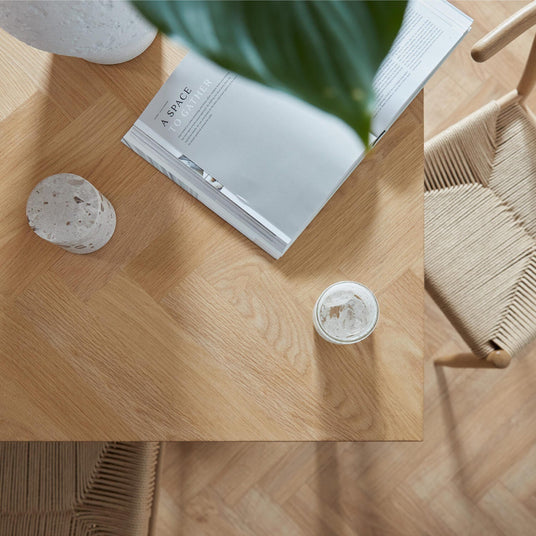 Top-down visning af hjørnet af et lyst egetræsbord med stol med flettet sæde, et opslået magasin med titlen "A SPACE TO GATHER", et terrazzo-objekt og et lille glas i blødt dagslys.