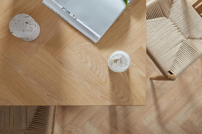 Top-down visning af hjørnet af et lyst egetræsbord med stol med flettet sæde, et opslået magasin med titlen "A SPACE TO GATHER", et terrazzo-objekt og et lille glas i blødt dagslys.