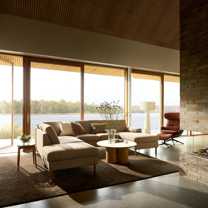 Rolig skandinavisk stue med beige hjørnesofa, brunt tæppe og rundt træbord foran store vinduer med udsigt til en sø; gulvlampe, loungestol og en vase med grene på bordet.