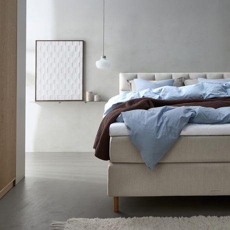 Lyst soveværelse med beige kontinentalseng redt med lyseblåt sengetøj og brun plaid, hvid pendel og billede på hylde.